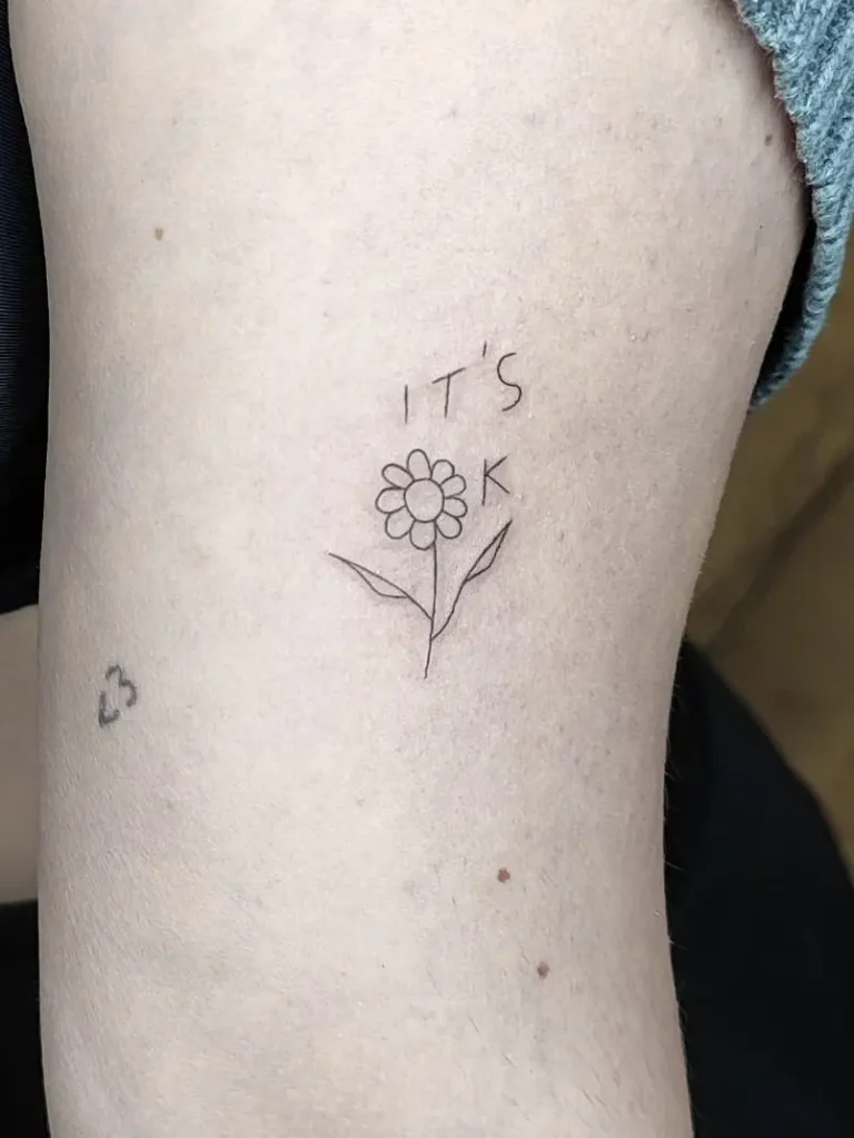 Tattoo de flor tipo margarita lineal con la frase It´s ok en el brazo
